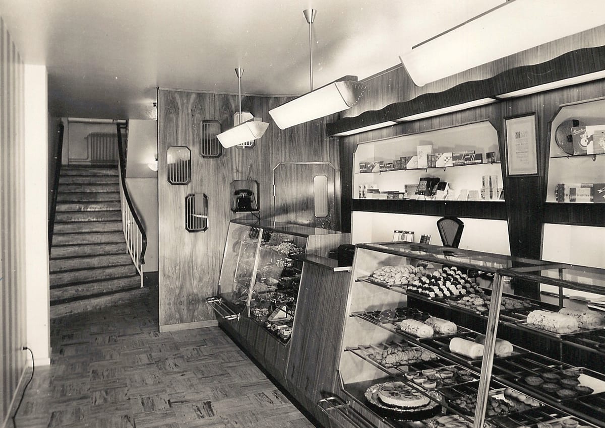 Svartvitt foto av Dackås konditori på Storgatan: en lång glasdisk full av bakverk och hyllor med chokladaskar